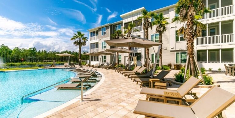 Modern apartment complex in the Wildlight master planned community, featuring a large outdoor swimming pool, lounge chairs, patio umbrellas, palm trees, and a clear blue sky.