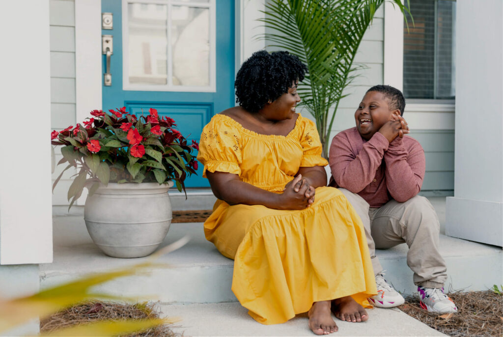 Two people sit on a front porch in the Wildlight Yulee Florida community; one wears a yellow dress, the other a maroon shirt and beige pants. They smile at each other beside a potted plant and blue door, enjoying homes in Nassau County Florida.