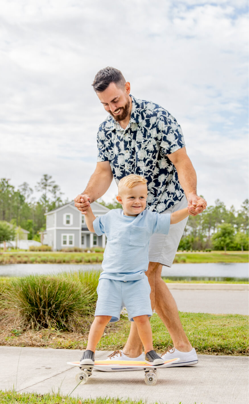 An adult helps a smiling young boy ride a skateboard on a sidewalk in the Wildlight Yulee Florida community, capturing a joyful moment in this vibrant Nassau County neighborhood on a cloudy day.