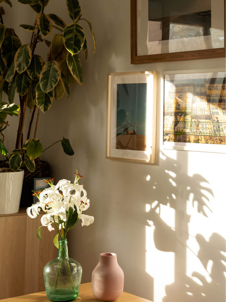 A sunlit corner in one of the stunning homes in Nassau County, Florida, features a green plant in a white pot, framed wall art, and a table holding a green glass vase with white lilies and a pink ceramic vase.