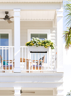 Second-story porch with white railing, ceiling fan, wicker chairs, and a flower box filled with white and yellow flowers; palm tree partially visible on the right—perfect for relaxing in the Wildlight master planned community.