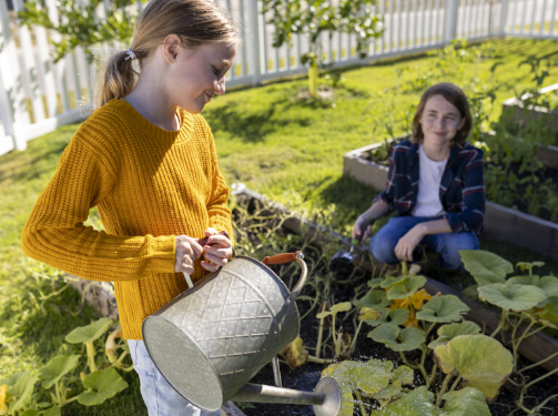 A girl in a yellow sweater waters plants in a garden while another person kneels nearby, watching her—capturing the peaceful spirit of the Wildlight Yulee Florida community.