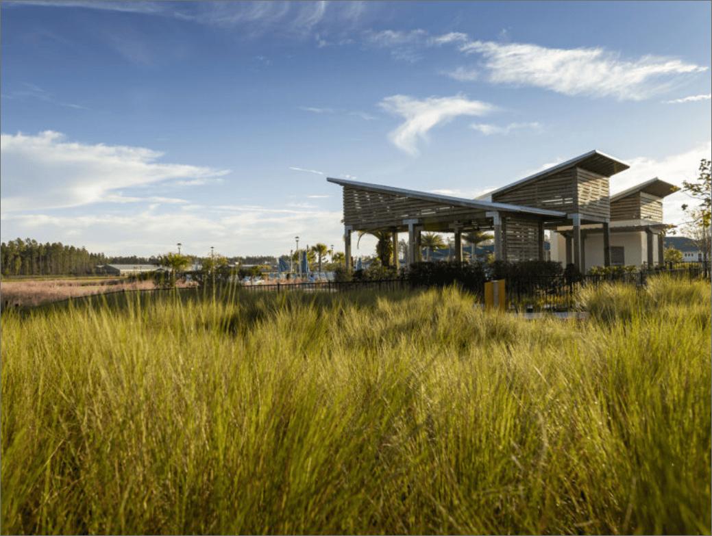 A modern open-air pavilion stands behind tall grass beneath a blue sky with scattered clouds in the landscaped outdoor area of the Wildlight Yulee Florida community.