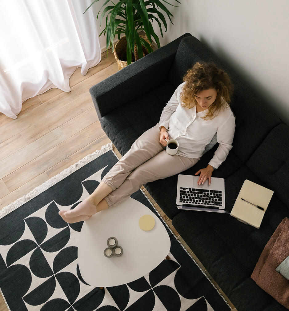 Woman sitting on a dark sofa with a laptop on her lap, holding a mug, next to a coffee table with a notebook, pen, and candles in a modern living room in the Wildlight Yulee Florida community.