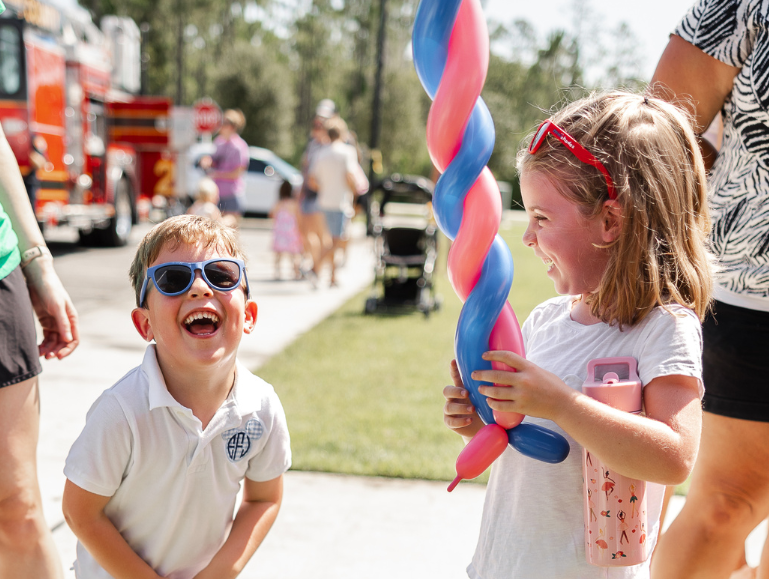 Two young children wearing sunglasses laugh outdoors in the Wildlight Yulee Florida community; one holds a twisted pink and blue balloon. Other people and emergency vehicles are visible in the background of this master planned community.