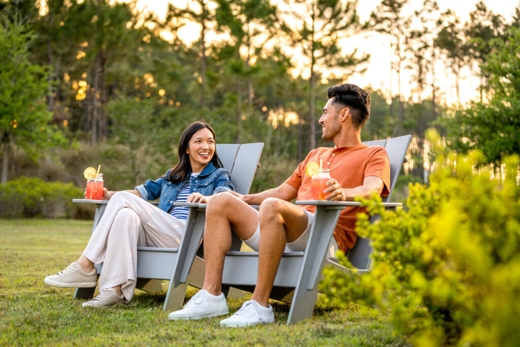 Two people sit in outdoor chairs on a lawn in the Wildlight Yulee Florida community, holding drinks with lemon slices, smiling and conversing, with trees in the background.