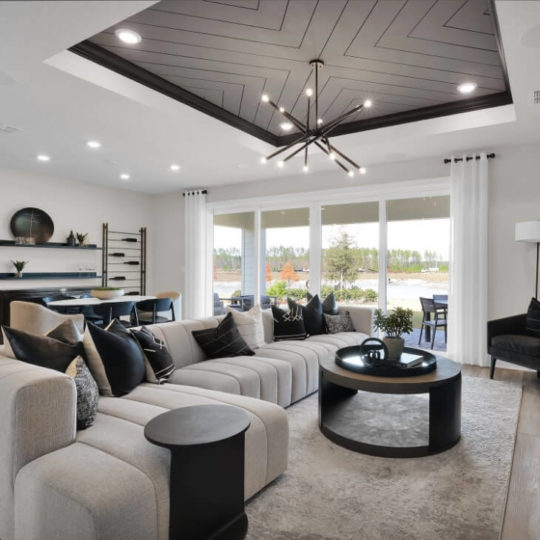 Modern living room with a large sectional sofa, round coffee tables, black and white decor, a geometric ceiling design, and large windows overlooking scenic views in the Wildlight Yulee Florida community.