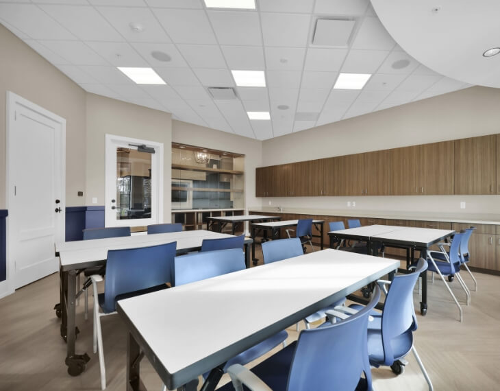 A modern classroom in the Wildlight Yulee Florida community features blue chairs, white tables, wood cabinets, and large windows in the background. The room is well-lit with ceiling lights.