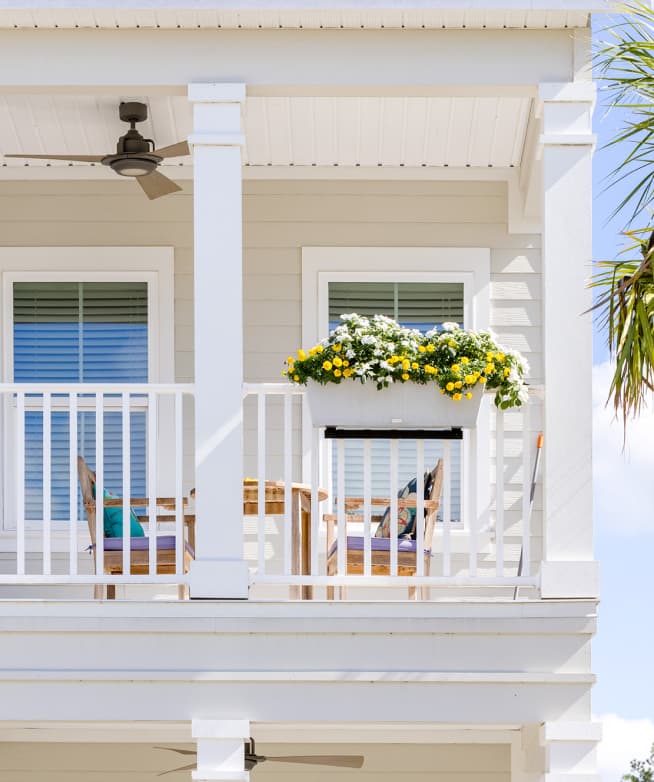 Second-story porch with white railing, ceiling fan, two chairs, table, and a planter box of yellow and white flowers. Enjoy relaxing views in the Wildlight master planned community, with a palm tree partially visible on the right side.