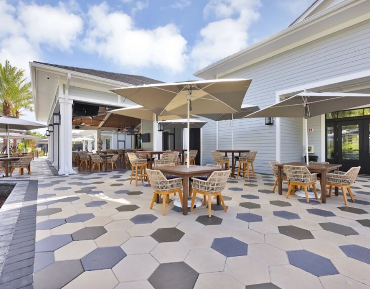 Outdoor patio with hexagonal tile flooring, wicker chairs, round wooden tables, and large umbrellas beside a light gray building under a partly cloudy sky in the heart of the Wildlight Yulee Florida community.