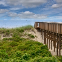 A wooden boardwalk extends over sand dunes with green vegetation in Wildlight Florida, leading toward a calm ocean under a partly cloudy sky—perfect for exploring near new homes in Wildlight FL.