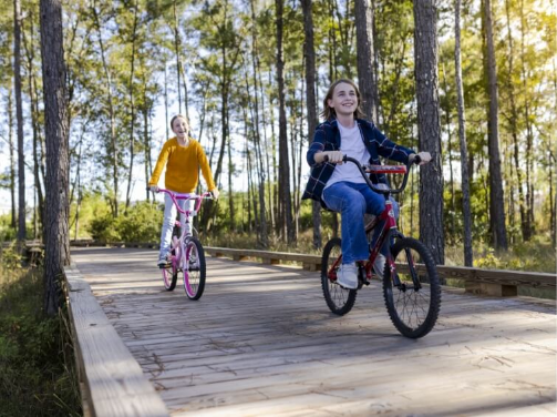 Two children ride bicycles on a wooden path through a forested area in Wildlight Florida, enjoying the sunny day within the vibrant Wildlight master planned community.
