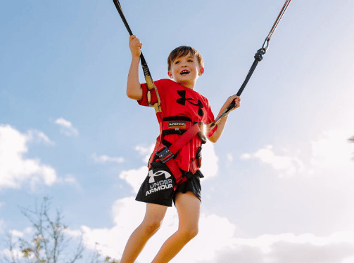 A child wearing a red Under Armour shirt and black shorts smiles while suspended in a harness on a bungee trampoline against a blue sky, enjoying the fun community spirit of Wildlight Florida.