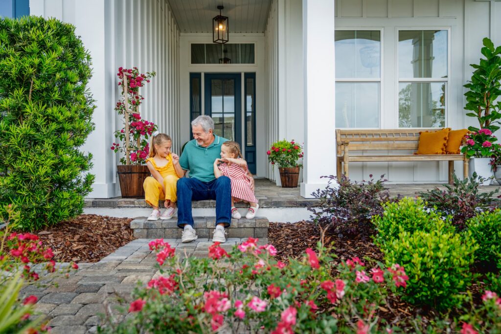 An older man sits on the front steps of a house with two young children, surrounded by flowers and greenery in the Wildlight Yulee Florida community.