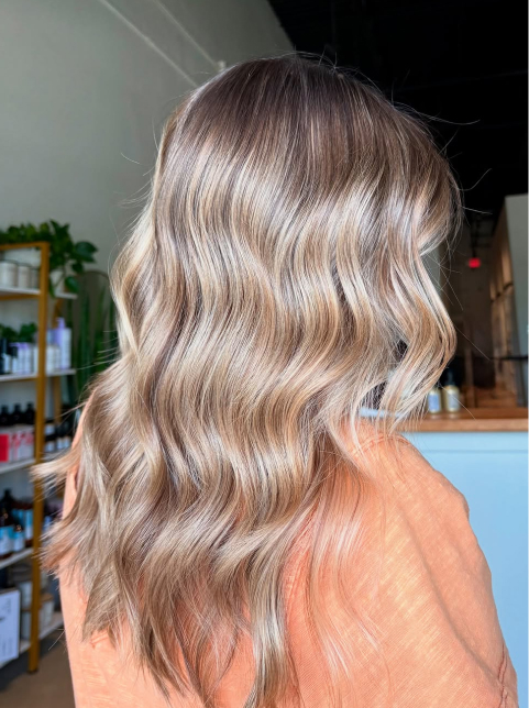 A person with long, wavy, light brown and blonde balayage hair is shown from the back in a salon setting, capturing the fresh style often seen among residents of the vibrant Wildlight Yulee Florida community.