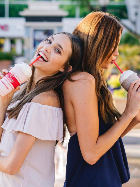Two young women stand back-to-back outdoors in the Wildlight Florida community, each drinking a milkshake with a red straw. One smiles at the camera, while the other looks down at her drink.