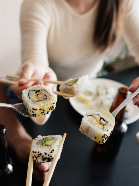 Four pieces of sushi are held up with chopsticks and forks by people gathered at a table, enjoying food and drinks—an inviting scene reminiscent of the lively community spirit found in new homes in Wildlight FL.
