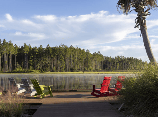 Six colorful Adirondack chairs sit on a wooden deck overlooking a calm lake in the serene Wildlight Florida community, with a forest backdrop under a blue sky.