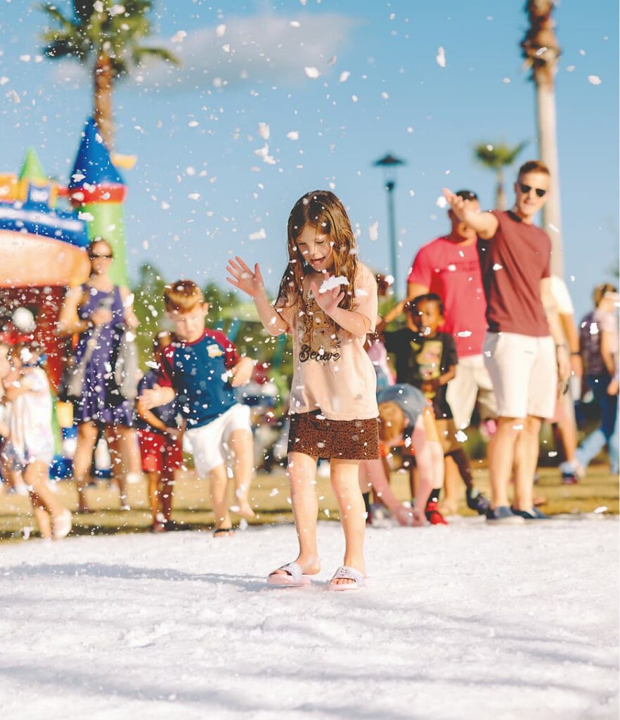 A young girl walks on artificial snow at a Wildlight Yulee Florida community event, with children and adults in summer clothing enjoying the scene around her.