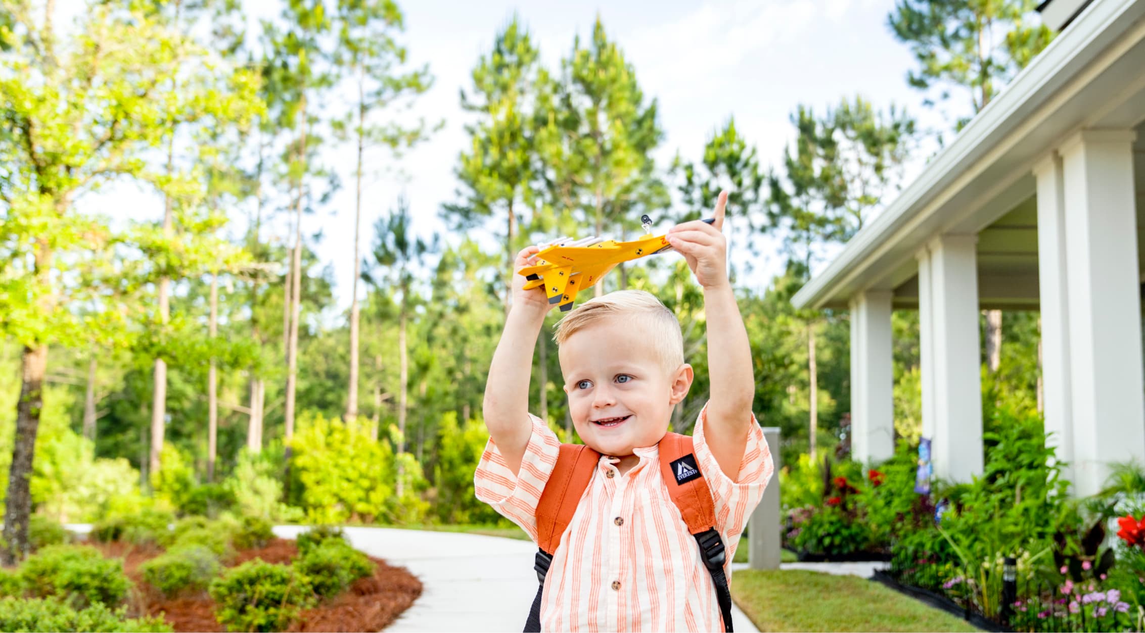 Young boy with a backpack holds up a toy dinosaur outside on a sunny day, standing near a path and a house surrounded by greenery in the Wildlight Yulee Florida community.