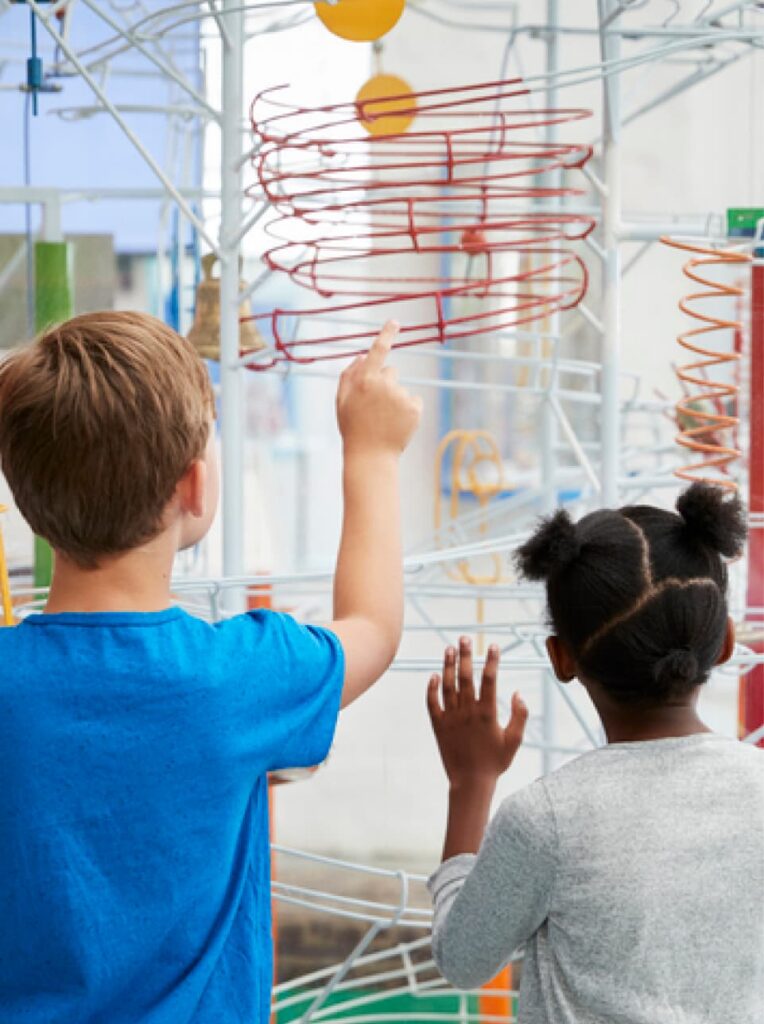 Two children stand side by side, exploring a large wire marble run exhibit at a discovery center—just one of many family-friendly activities near the new homes in Wildlight FL.