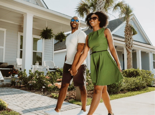 A couple holding hands and walking on a sidewalk in front of a modern house with a porch, palm trees, and landscaped garden in the vibrant Wildlight Yulee Florida community.