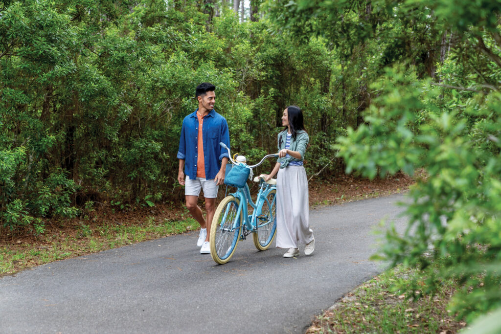 A man and a woman walk side by side on a paved path in a wooded area within the Wildlight Yulee Florida community, with the woman holding a blue bicycle.