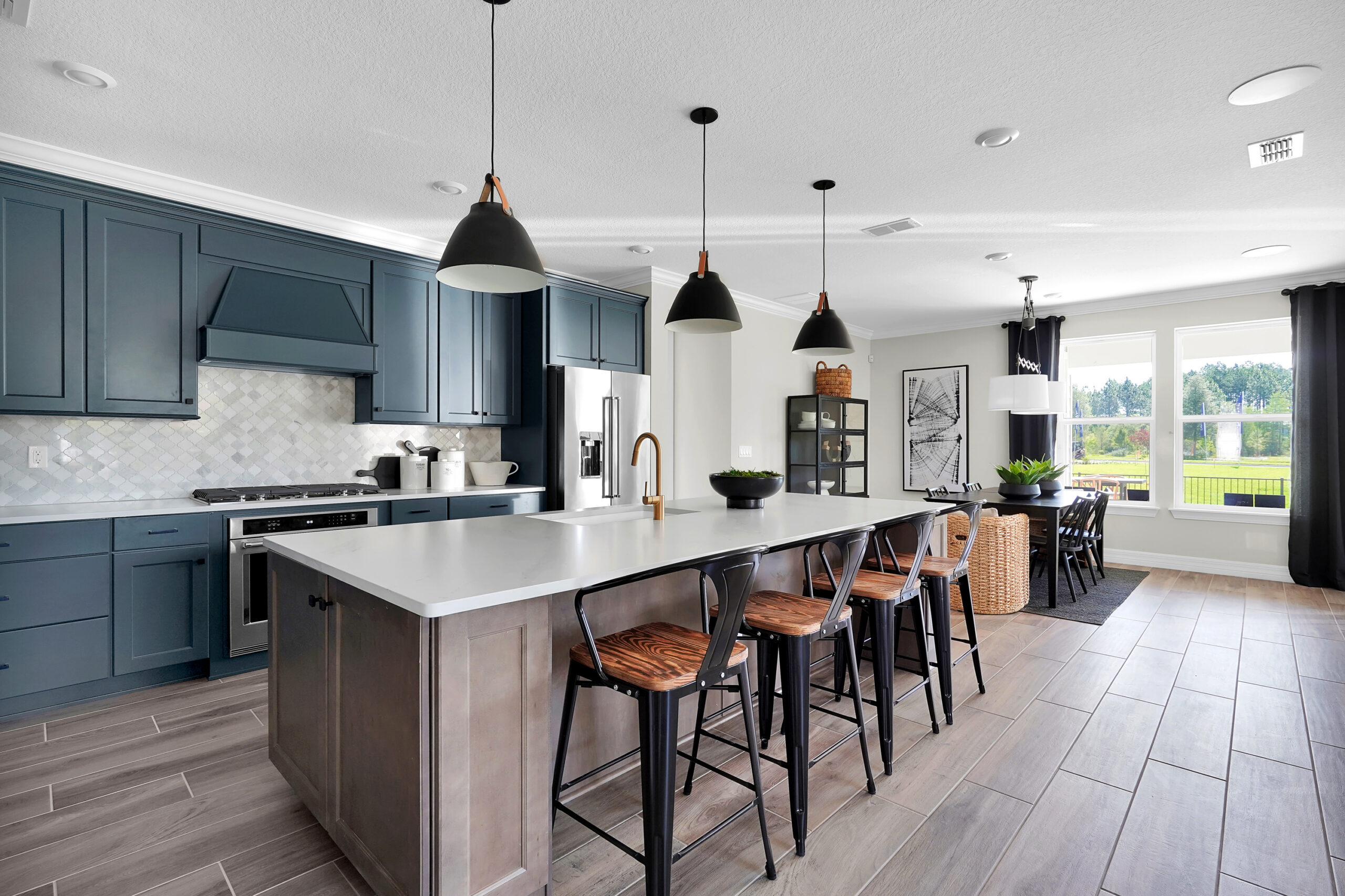 Modern kitchen with blue cabinets, stainless steel appliances, a large island with seating, pendant lights, and a dining area by large windows—perfect for enjoying the green views in the Wildlight Yulee Florida community.