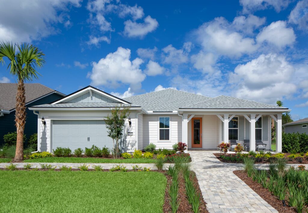 Single-story modern house with a light gray exterior, front porch, attached garage, and landscaped yard in the Wildlight master planned community. Features a palm tree and paved walkway under a blue sky with clouds—discover new homes in Wildlight FL.