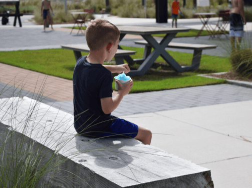 A young boy sits on a wooden bench outdoors in Wildlight Florida, holding a cup of blue shaved ice, with picnic tables and people in the background of this vibrant master planned community.