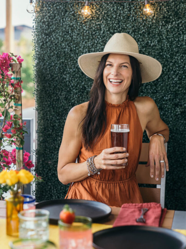 A woman in a wide-brimmed hat and orange dress sits at an outdoor table, smiling and holding a drink. The scene, reminiscent of homes in Nassau County Florida, is set with plates, flowers, and a strawberry.