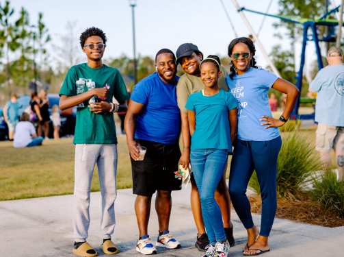 A group of five people, four adults and one child, stand smiling together outdoors in a park on a sunny day in the Wildlight Yulee Florida community.