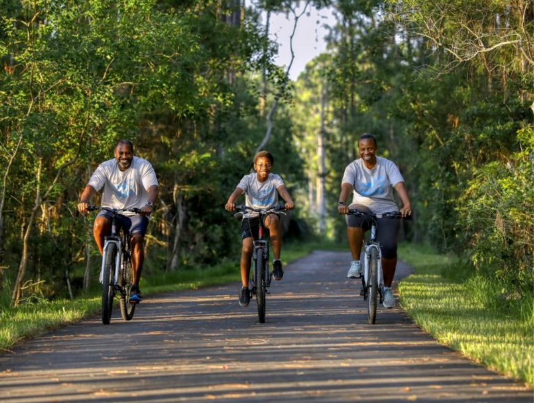 Three people ride bicycles on a paved path through a green, wooded area in the Wildlight Yulee Florida community on a sunny day.