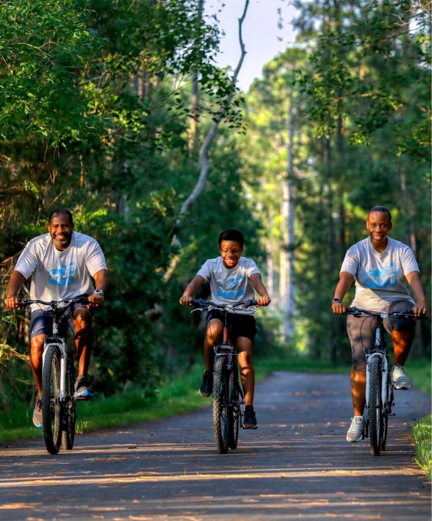 Three people ride bicycles on a paved path through a wooded area in Wildlight Florida, all wearing gray shirts and smiling, with sunlight filtering through the trees.