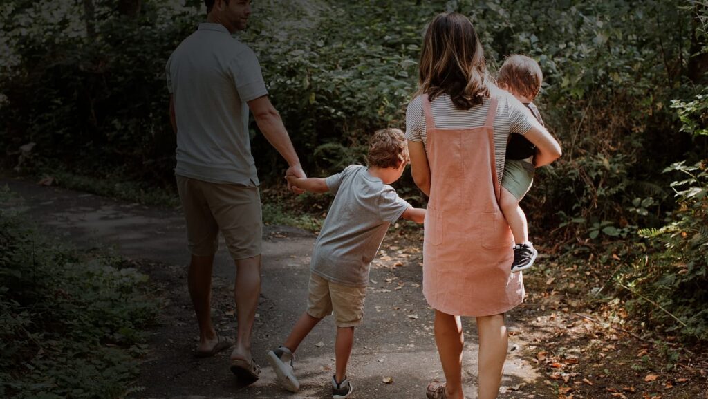 Two adults and two children walk together on a path through a wooded area in the Wildlight Yulee Florida community; one adult carries a small child while holding hands with another child.