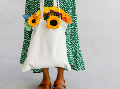 A person wearing a green polka dot dress and brown sandals holds a white tote bag filled with sunflowers, enjoying the vibrant atmosphere of the Wildlight Yulee Florida community.