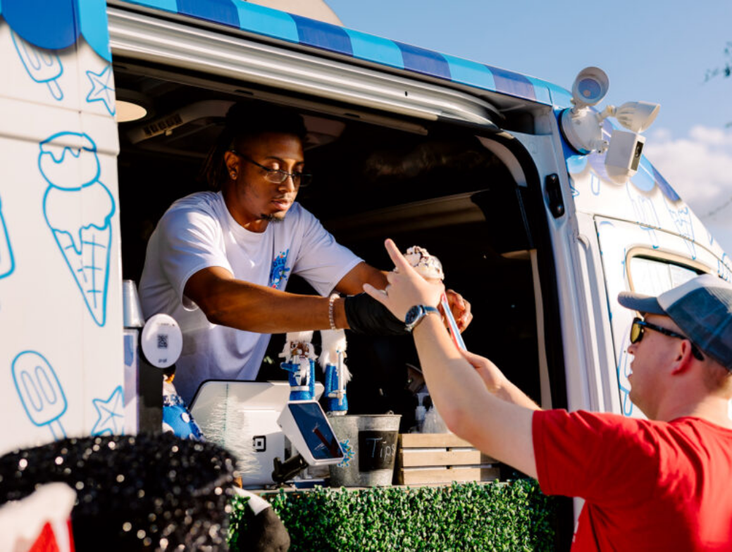 A vendor hands an ice cream cone to a customer at the window of an ice cream truck decorated with blue and white graphics in the heart of Wildlight Florida, a vibrant master planned community.