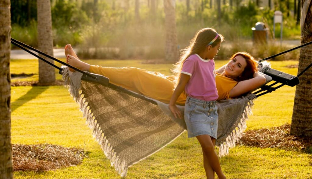A woman lies on a hammock while a young girl stands beside her in a grassy outdoor area with trees in the background, capturing the relaxing lifestyle found in the Wildlight Yulee Florida community.