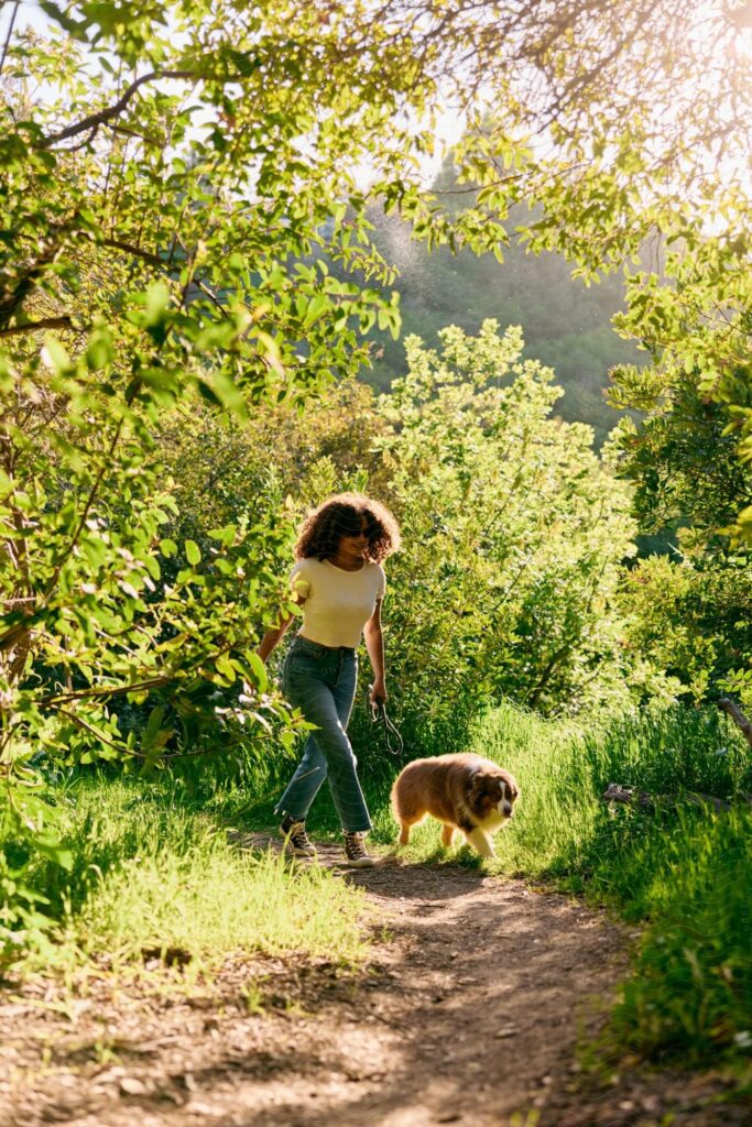 A woman walks a dog along a sunlit dirt path surrounded by green trees and foliage in the Wildlight Yulee Florida community.