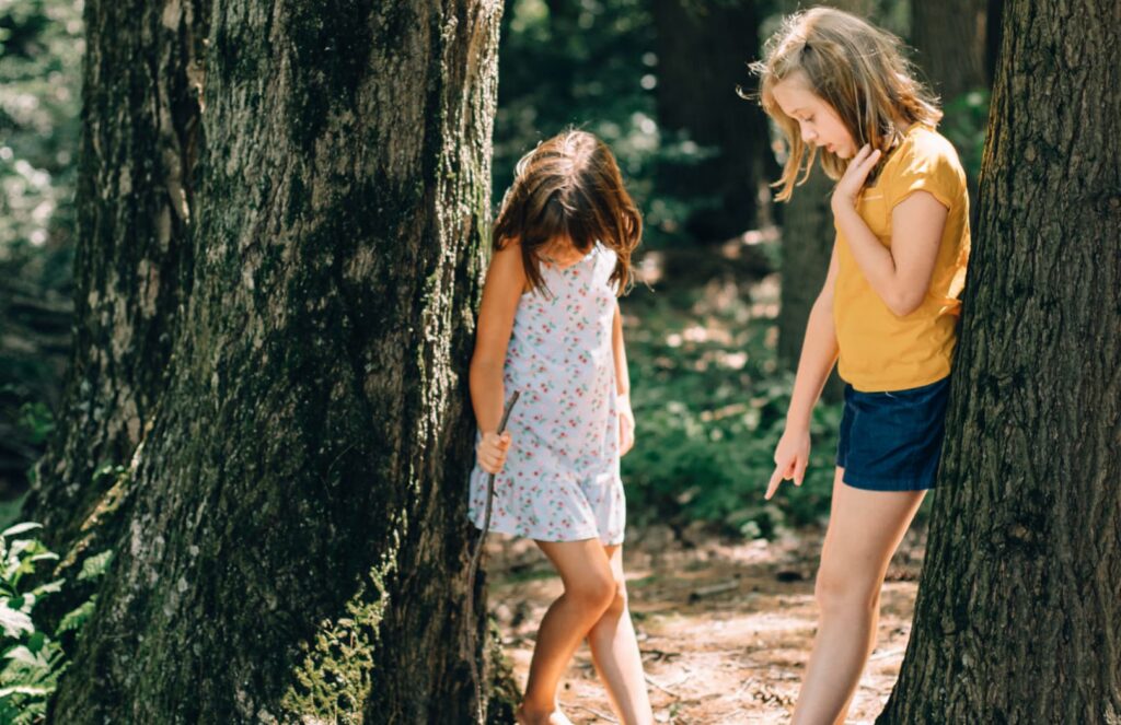 Two young girls stand by large trees in a forest within the Wildlight Yulee Florida community. One girl in a yellow shirt and blue shorts points at something on the ground while the other looks down, exploring nature together.