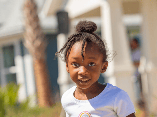 A young girl with braided hair and a white shirt stands outside, looking at the camera with a slight smile. Blurred buildings and greenery hint at charming new homes in Wildlight FL in the background.