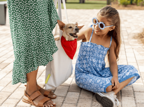 A woman stands beside a girl sitting on a brick path; a small dog wearing a red bandana sits in the woman’s tote bag, capturing the welcoming spirit of the Wildlight Yulee Florida community.