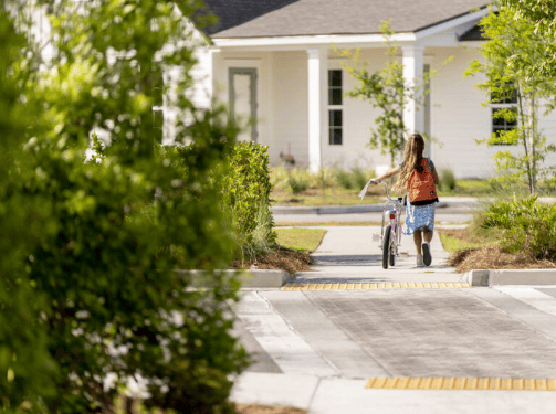 A girl with a backpack walks her bicycle along a sidewalk toward a white house, surrounded by greenery in the Wildlight Florida community on a sunny day.