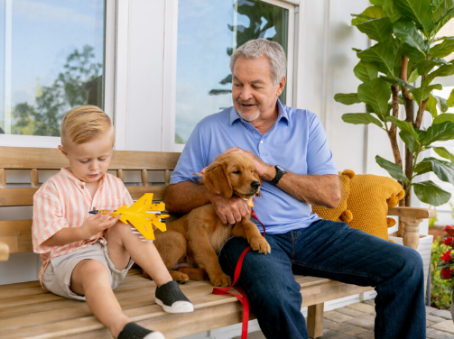 A man sits on a wooden bench with a young boy playing with a toy airplane and a golden retriever puppy beside them, enjoying the family-friendly atmosphere of the Wildlight Yulee Florida community.