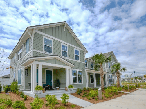 Two-story modern suburban house with green siding, white trim, front porch, and landscaped yard with palm trees under a partly cloudy sky—one of the stunning new homes in Wildlight FL.