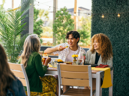 Three people sit at an outdoor table with meals and drinks, talking and smiling under string lights, surrounded by greenery—enjoying the relaxed lifestyle of the Wildlight master planned community.
