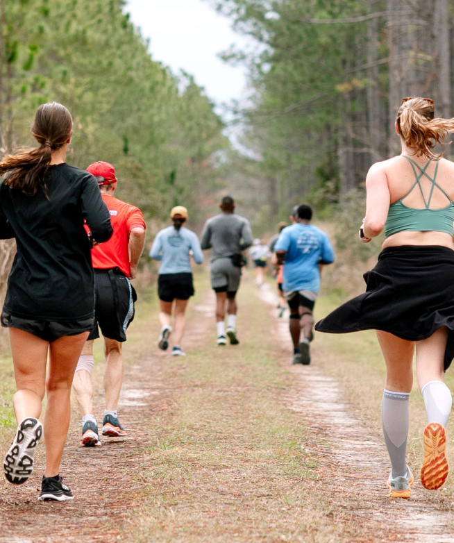 A group of people run on a dirt trail through a forested area in Wildlight Florida, dressed in athletic wear, with trees lining both sides of the path in this vibrant master planned community.