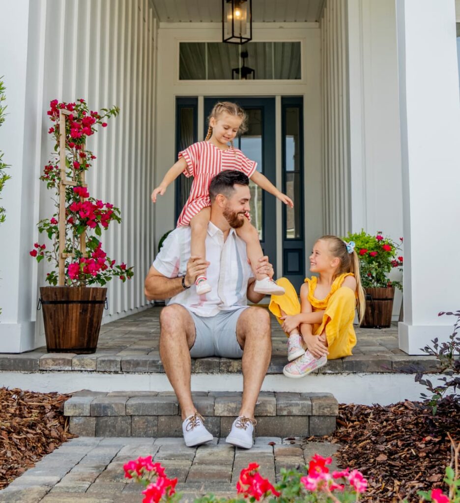An adult and two children sit on the front steps of their new home in Wildlight FL, with potted flowers nearby. One child sits on the adult’s shoulders while the other smiles from the step—a joyful moment in this vibrant master planned community.