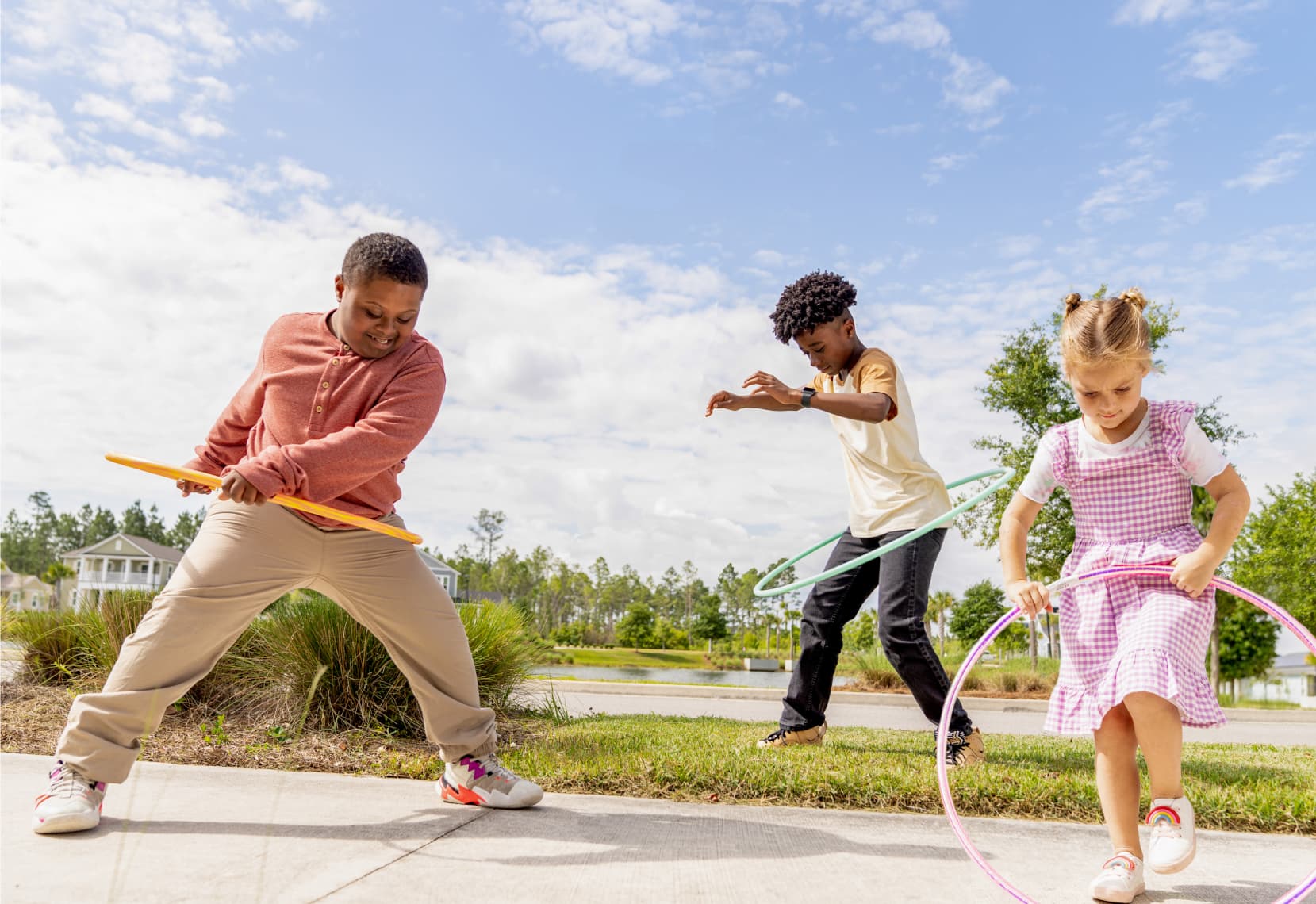 Three children play with hula hoops on a sunny day in the Wildlight Yulee Florida community, standing on a sidewalk surrounded by grass, trees, and charming new homes in Wildlight FL.