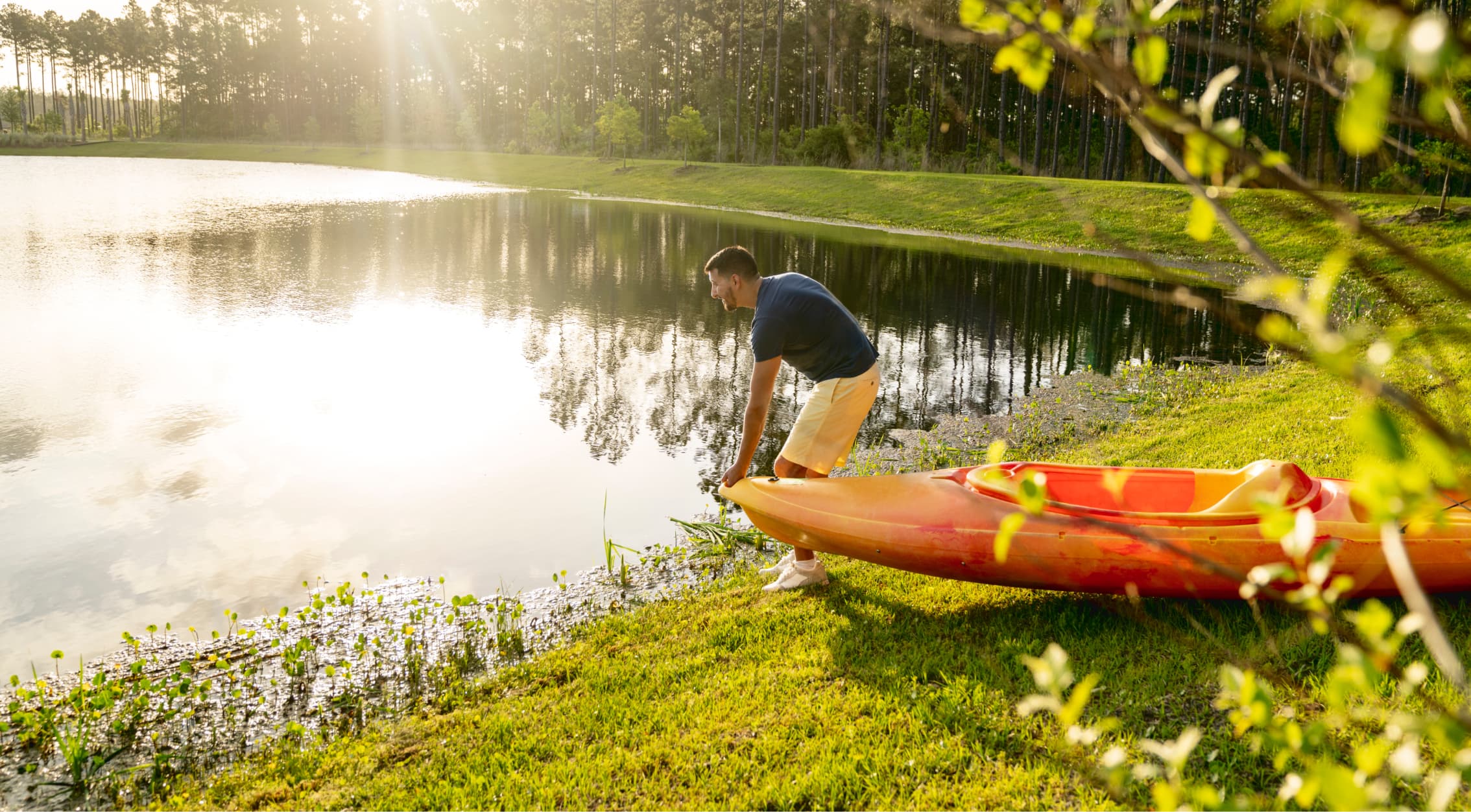 A man stands on the grassy shore of a lake in Wildlight Florida, preparing to launch a red kayak into the water, with trees and sunlight illuminating the background near new homes in Nassau County Florida.